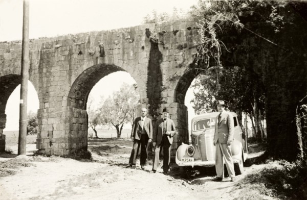 Visiting the aqueduct near Acre. Three men in suits stand beside a car beneath the arches of the