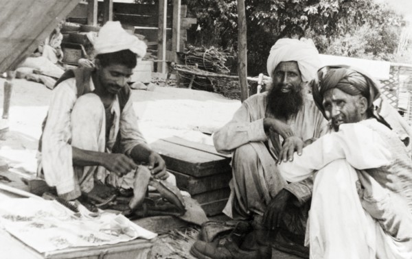 A visit to the cobbler. Two elderly men visit a cobbler who squats on the ground, mending boots in