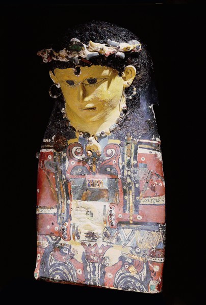 Mummy case portrait mask of a young woman with curled hair, with glass beaded bronze hoop earrings, inset beads, and glass inlay on chest depicting two orange ankh signs flanked by four Seth headed wa...