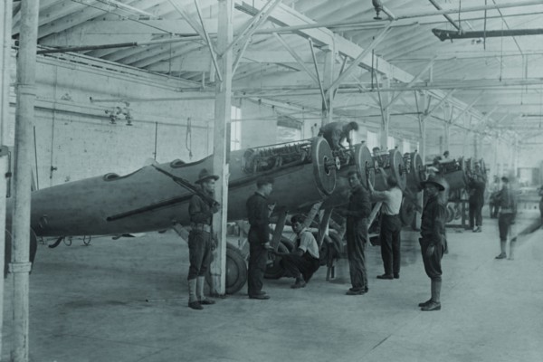 A line of airplanes is being assembled under the watchful eye of a gun-toting soldier as they are being completed to be sent to the European war; the factory is at College Point, Queens 1917