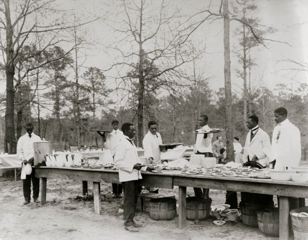 African American men preparing to serve a meal in an outdoor setting among trees 1900