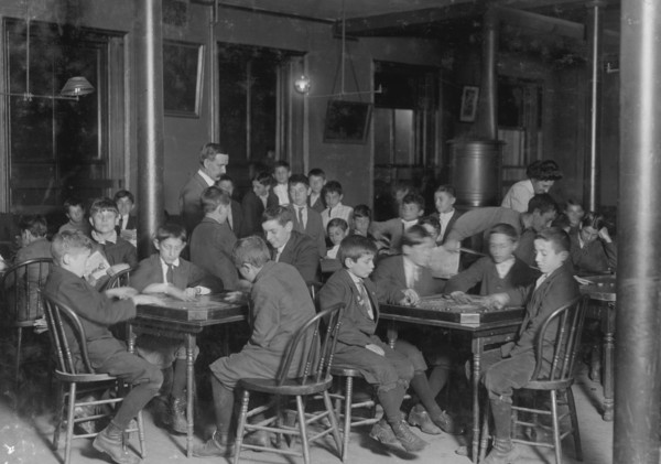 In the Newsboys Reading Room. Boys seated at tables playing gamers. 1909