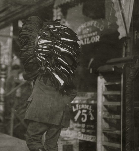 Man loaded down with heavy bundle of vests that he had been carrying for many blocks, stopping to rest, occasionally, when he would drop the bundle down onto whatever boxes or railings were available,...