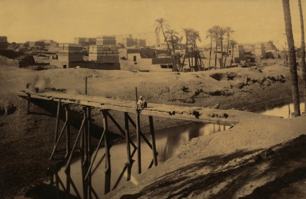 Man seated on wooden bridge over canal, ruins and buildings in background 1880