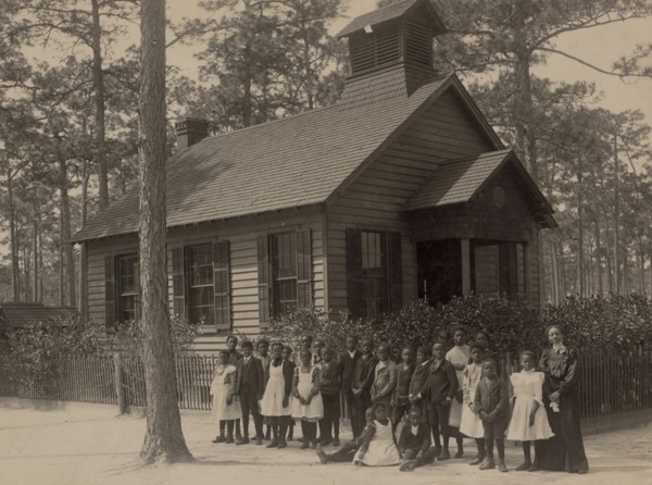 African American school children posed with their teacher outside a school, possibly in South Carolina
