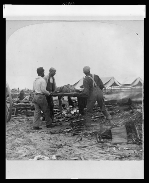 Galveston, Texas, 1900 - African-American men carrying a body on a stretcher, surrounded by wreckage of the hurricane and flood. Stereograph.