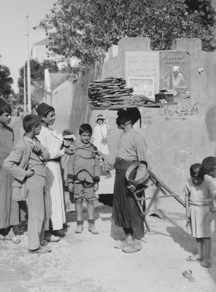 A man, street vendor, selling cakes in a Beirut street  to a crowd of young boys, man has cakes on top of his head ca. 1900
