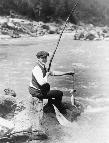 Man seated on rock, with two fish on line, by stream, possibly a river, Washington, D.C. area ca. 1909