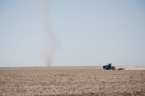 Farmer plowing a field on the Eastern Colorado plains, tractor facing left and driving toward a natural phenomena called a dust devil
