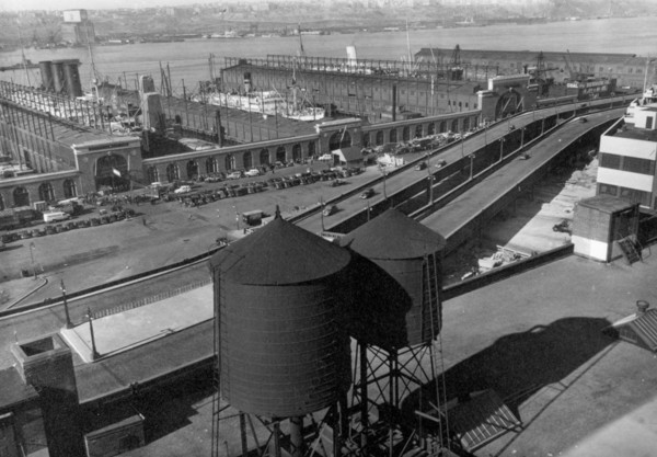 Eight cars visible on Westside highway, beyond water towers in foreground, piers have entrances on street, ships visible at dock ca. 1937