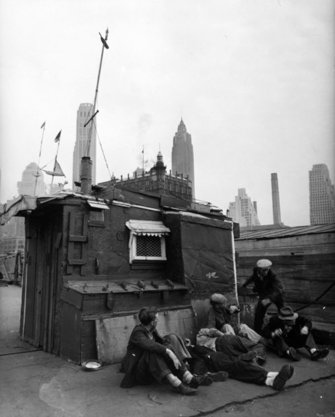 Men in front of shelter, decorated with flags on masts, ship-carvings, window with awning. One man seems to have just hit another ca. 1938