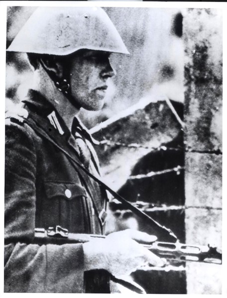 A Helmeted East German Policeman, Holding a Sten Gun Stands Guard Near a Barbed Wire Barricade at