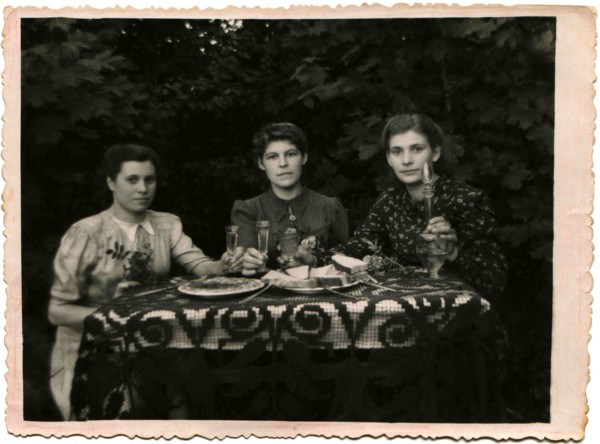Three young women holding glasses of alcohol, sitting at a table covered with in garden.