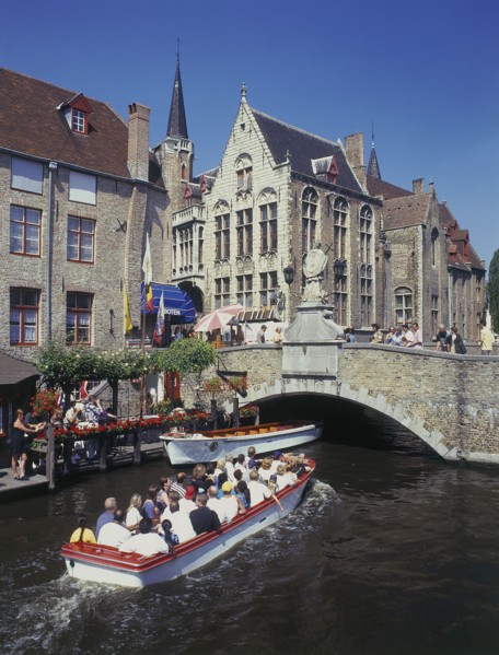 BELGIUM, West Flanders, Bruges, Tourists on boat trips on a canal with old bridge and houses behind