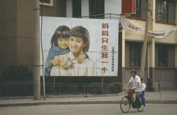 CHINA, Shandong, Jinan, Man with child passenger cycling past birth control poster with image of