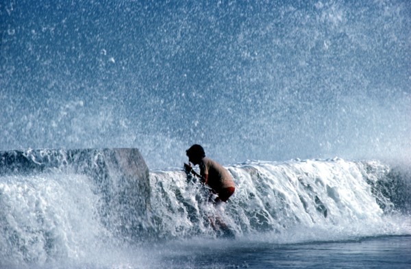 CUBA, Havana, Malecon, Young boy standing on pavement with waves crashing over the sea wall