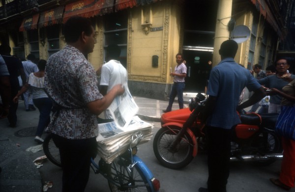 CUBA, Havana, Newspaper vendor selling goods from a bicycle seat in the street with passing traffic
