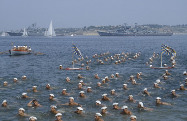 RUSSIA, Sevastopol, Navy Day. Sailors swimming in the sea in formation with battleships behind.
