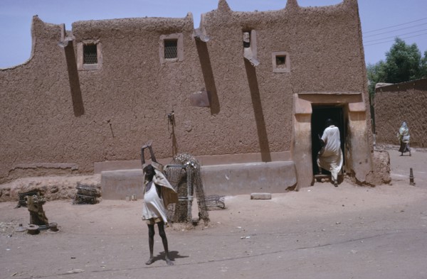 NIGERIA, Kano, Traditional mud Hausa dwelling with child standing with up-stretched arms outside