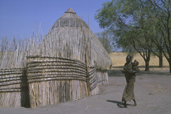 SUDAN, South, Woman and child in Shilluk village walking past thatched hut and enclosure.