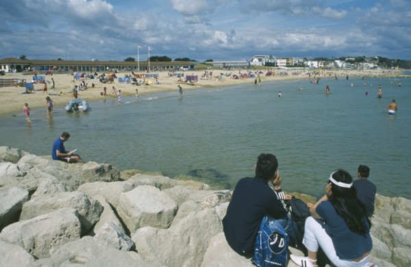 ENGLAND, Dorset, Sandbanks, People sat on rocks looking across water and busy sandy beachfront