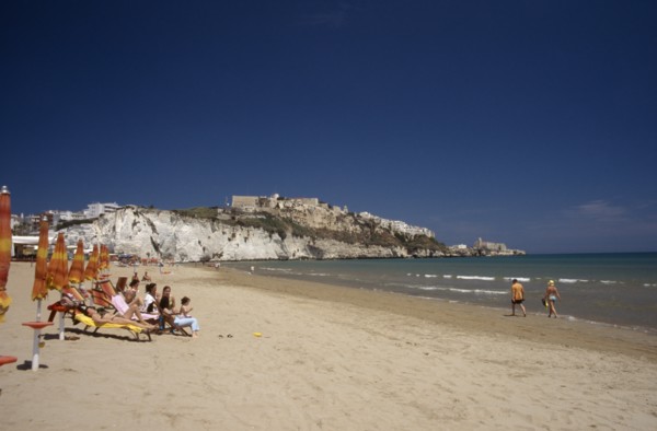 ITALY, Puglia, Foggia, Vieste.  Line of sunbathers on stretch of sandy beach overlooked by castle
