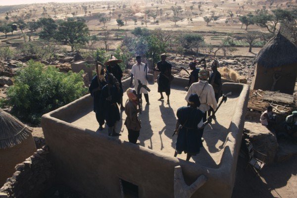 20086075 MALI People Dogon Members of ancient Dogon society performing rooftop dance holding carved staffs with view over surrounding village and landscape.