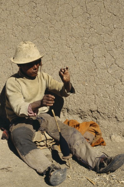 BOLIVIA, Chukiuta, Aymara man sitting against a wall with a spindle of thread. Near Sucre