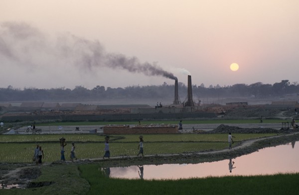 BANGLADESH, Savar, Labourers returning to brick kiln through paddy fields with chimneys releasing