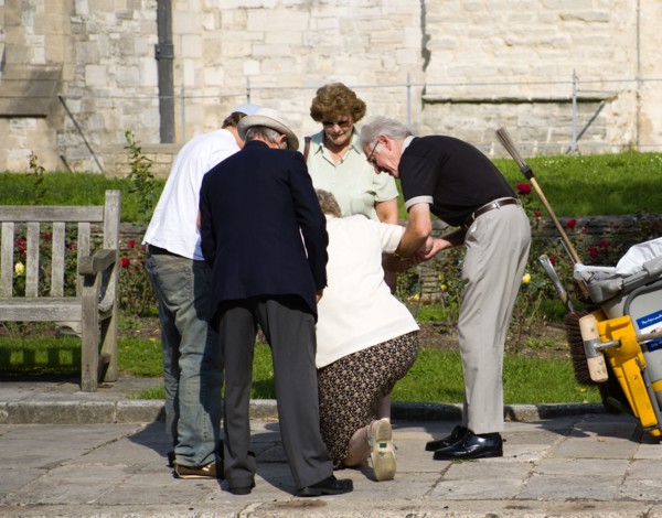 People helping an elderly lady to stand up after she has fallen on the pavement sidewalk outside