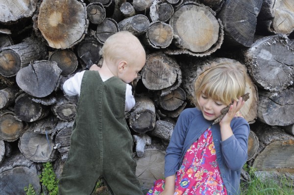 5 year old Eva and Oscar aged two playing beside a log pile with Eva using bark as pretend mobile phone.