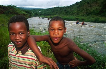 two boys on bank of river look into camera, paddlers behind on r