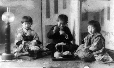 Three Japanese children seated on cushions on the floor,  eating noodles with chopsticks
