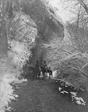 Two Crow Indians on horseback in shallow stream flanked by snow-covered trees