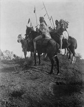Four Crow men on horseback holding feathered spears