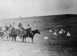 Four Atsina Indians on horseback overlooking tepees in valley beyond