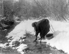 Apsaroke woman standing in snow scooping water from a stream with a can