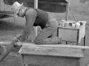White migrant worker sawing wood for stakes to be used in setting up tent home, near Harlingen, Texas by Russell Lee, 1903-1986, dated 19390101.