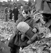 Abandoned boy holding a stuffed toy animal amid ruins following German aerial bombing of London during the Blitz of World War two