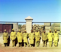 Group of labourers standing, poised, in front of a chain-link fence, with sacks piled behind fence.