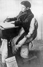 Boy seated at desk in sleeping bag 1912