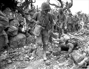Photograph of Okinawa U.S. Marines pass a dead Japanese soldier in a destroyed village