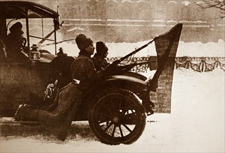 Photograph of soldiers riding on the mud flaps of a car with red flags attached to their guns in Petrograd