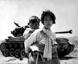 With her brother on her back a war weary Korean girl tiredly trudges by a stalled tank, at Haengju, Korea