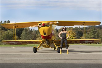 Young woman with sunglasses in overall and boots posing in front of double-decker airplane