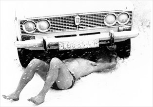 Man lies naked under a car in the snow ca. 1970s