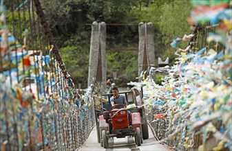 Farmer drives with a tractor over a suspension bridge decorated with prayer flags