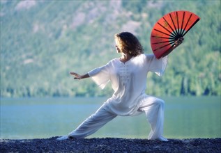 Young woman practicing Tai Chi Chuan with a red fan at mountain lake shore in the nature