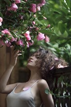 Artistic sensual portrait of a young beautiful woman standing outdoors by the garden fence under a blooming camellia tree touching its pink flowers