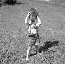 Child stands barefoot in a meadow and drinks water from a wooden-coated vessel